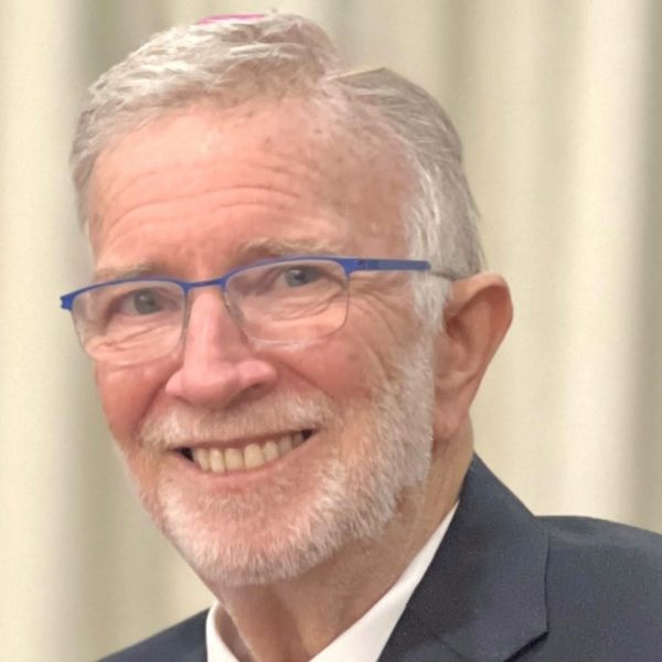 An older man with short gray hair, a neatly trimmed white beard, and blue glasses smiles warmly. He is wearing a dark suit jacket and a white shirt, with a light-colored background behind him.