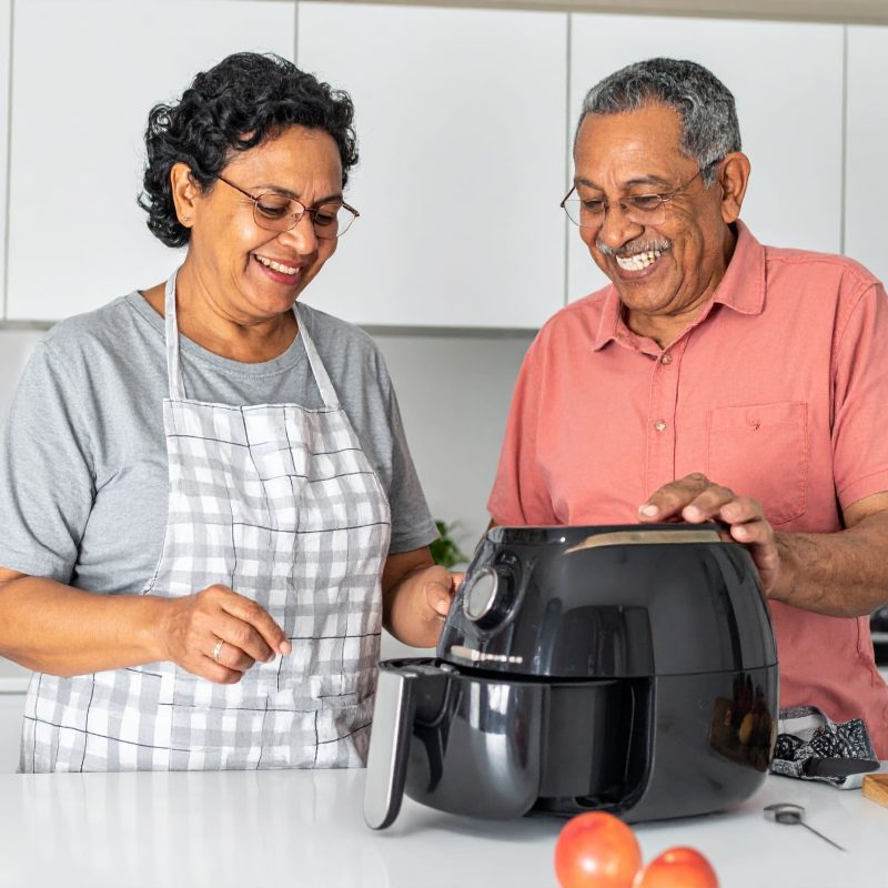 Seniors using an air fryer A smiling senior couple in a modern kitchen happily preparing food together with a black air fryer on the counter, surrounded by fresh fruits and vegetables.
